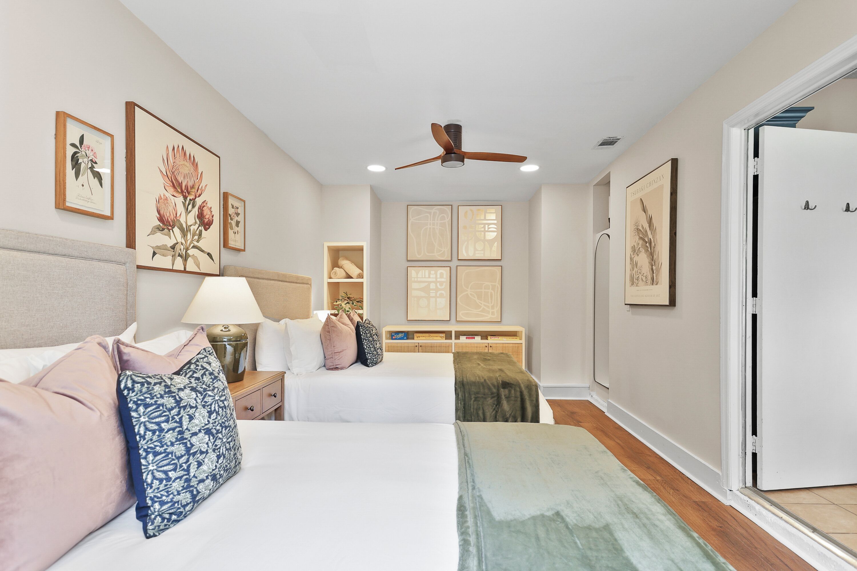 A hotel room with two beds, a ceiling fan, artwork on the walls, a bedside table with a lamp, and a wooden floor.