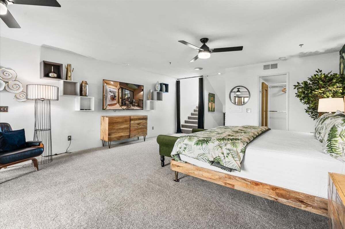 A bedroom with a bed, a dresser, a chair, a lamp, a ceiling fan, a staircase, and a door.
