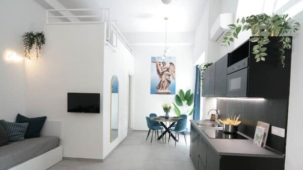 Una cocina moderna con armarios negros, un comedor con sillas azules y una sala de estar con sofá gris.