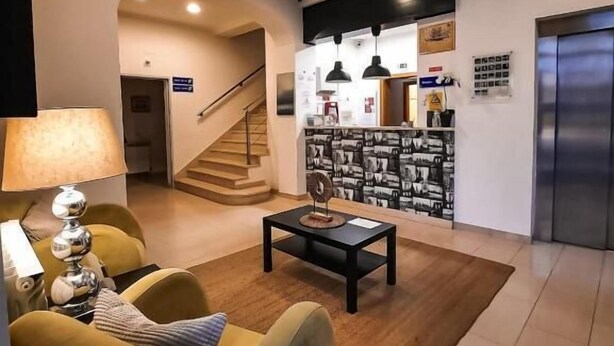 A modern living room with a sofa, coffee table, and lamp. There is a staircase leading to another level, a wall with framed pictures, and an elevator. - Hotel Vila Verde (Castro Verde)