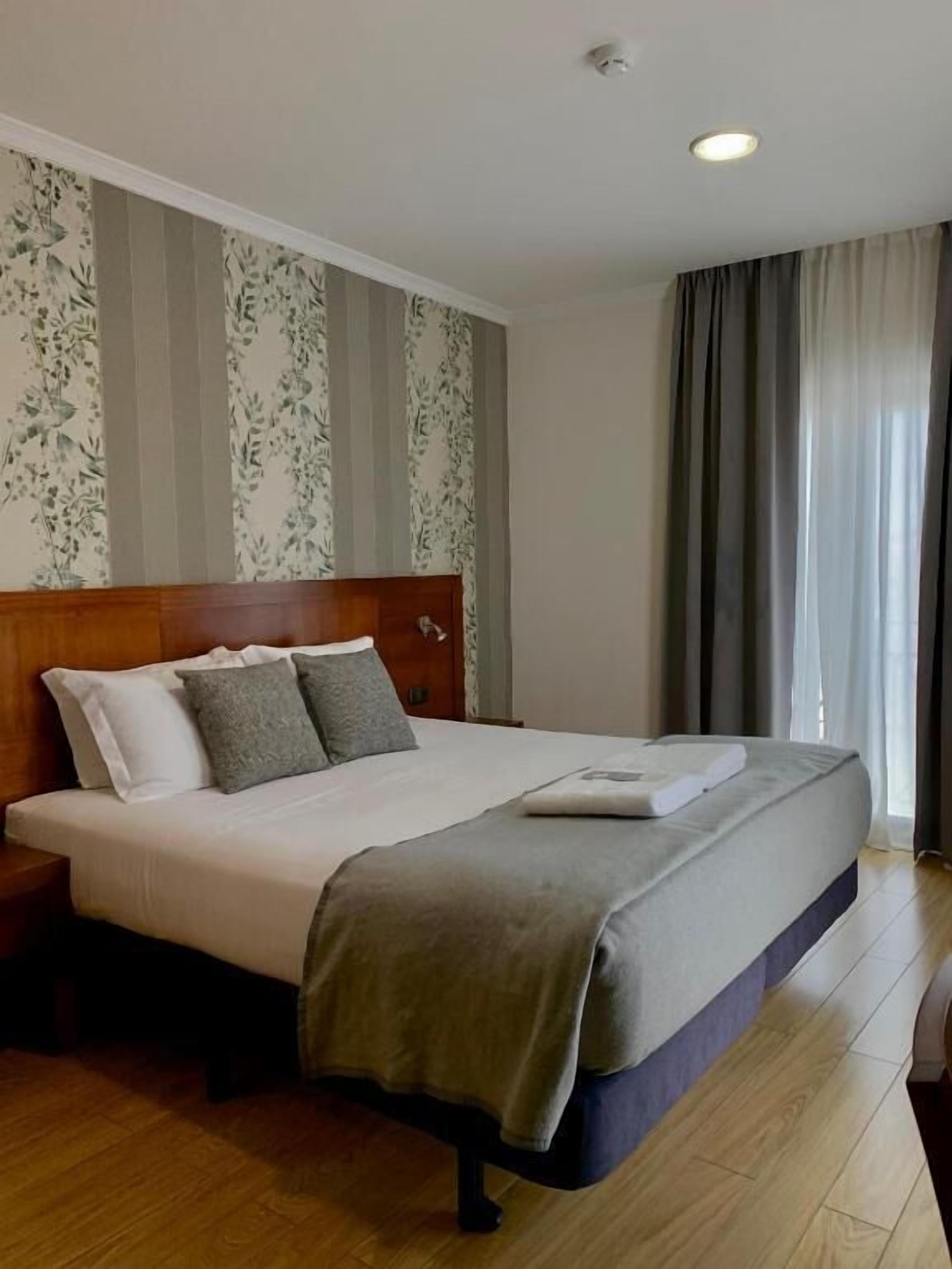 Habitación de hotel con una cama grande, cabecero de madera, papel tapiz floral y vistas al exterior a través de una ventana con cortinas.