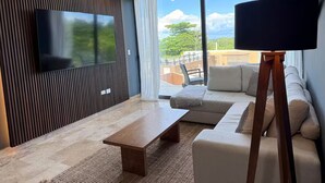 A modern living room with a flat-screen TV mounted on a wooden panel wall, a wooden coffee table, a beige sofa with patterned cushions, and a large window offering a view of greenery and a building.