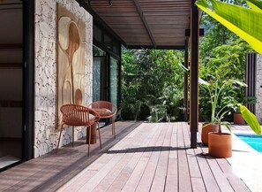Um pátio externo com piso de madeira, duas cadeiras de vime, uma parede de pedra com escultura em relevo e uma piscina ao fundo.