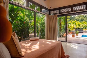 A bedroom with a bed, large windows, and a view of a pool and greenery.