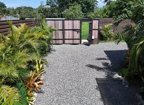 Um caminho de cascalho leva a um portão de madeira com fecho de metal, cercado por plantas tropicais.
