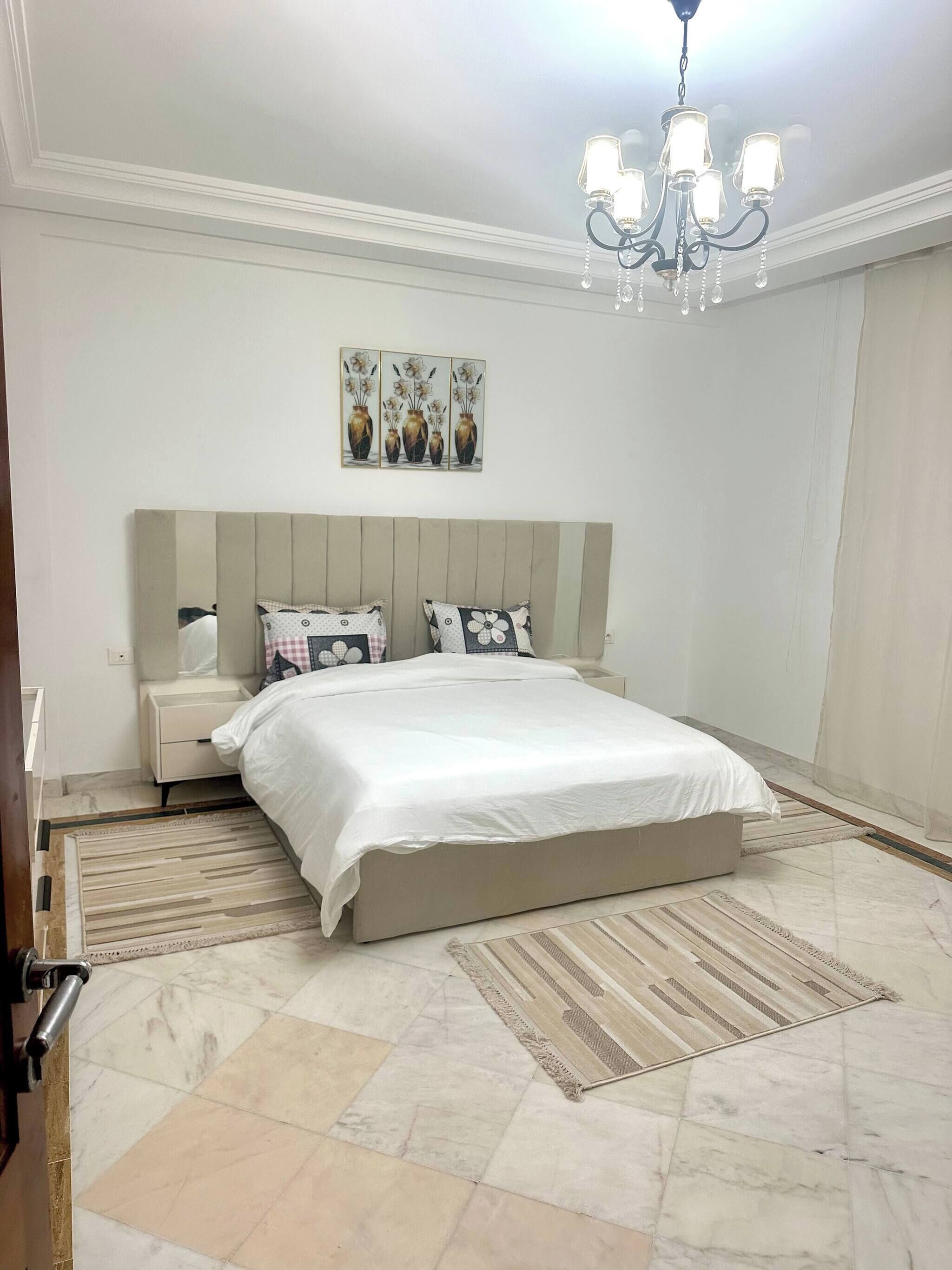 A bedroom with a large bed, a nightstand, a chandelier, and a painting on the wall.