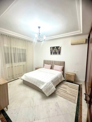 A bedroom with a bed, bedside tables, a chandelier, an air conditioner, and a window with sheer curtains. - Luxury Stay With Panoramic Sea Views, Tourist Zone (Sousse)