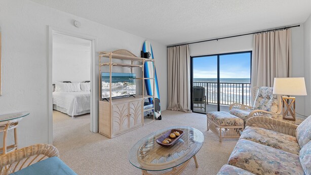 A hotel room with a bed, a glass table with a tray of snacks, a wicker sofa, a chair, a balcony with a view of the ocean, and a large window with curtains.