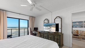 A hotel room with a large bed, a television, a desk with a chair, and a view of the beach.
