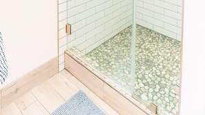 Ducha con cerramiento de vidrio, azulejos blancos y verdes, piso de madera y alfombra azul.