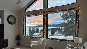 A living room with a large window overlooking a snowy landscape and mountains, decorated with festive lights.