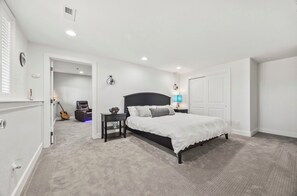 A bedroom with a large bed, a nightstand, a lamp, a guitar, and a door leading to another room.