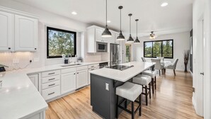 A modern kitchen with white cabinetry, a central island with bar stools, pendant lighting, and large windows offering views of the outdoors.