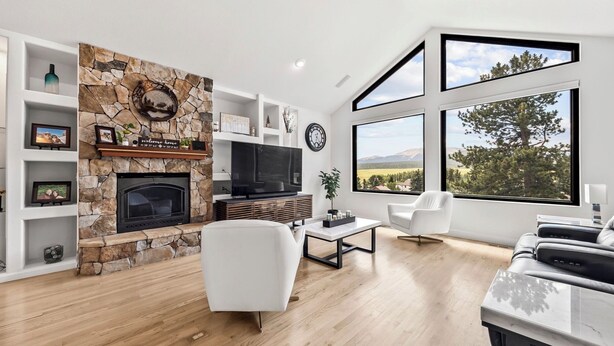 A modern living room with a stone fireplace, a flat-screen TV, white furniture, and large windows offering a view of trees and mountains.
