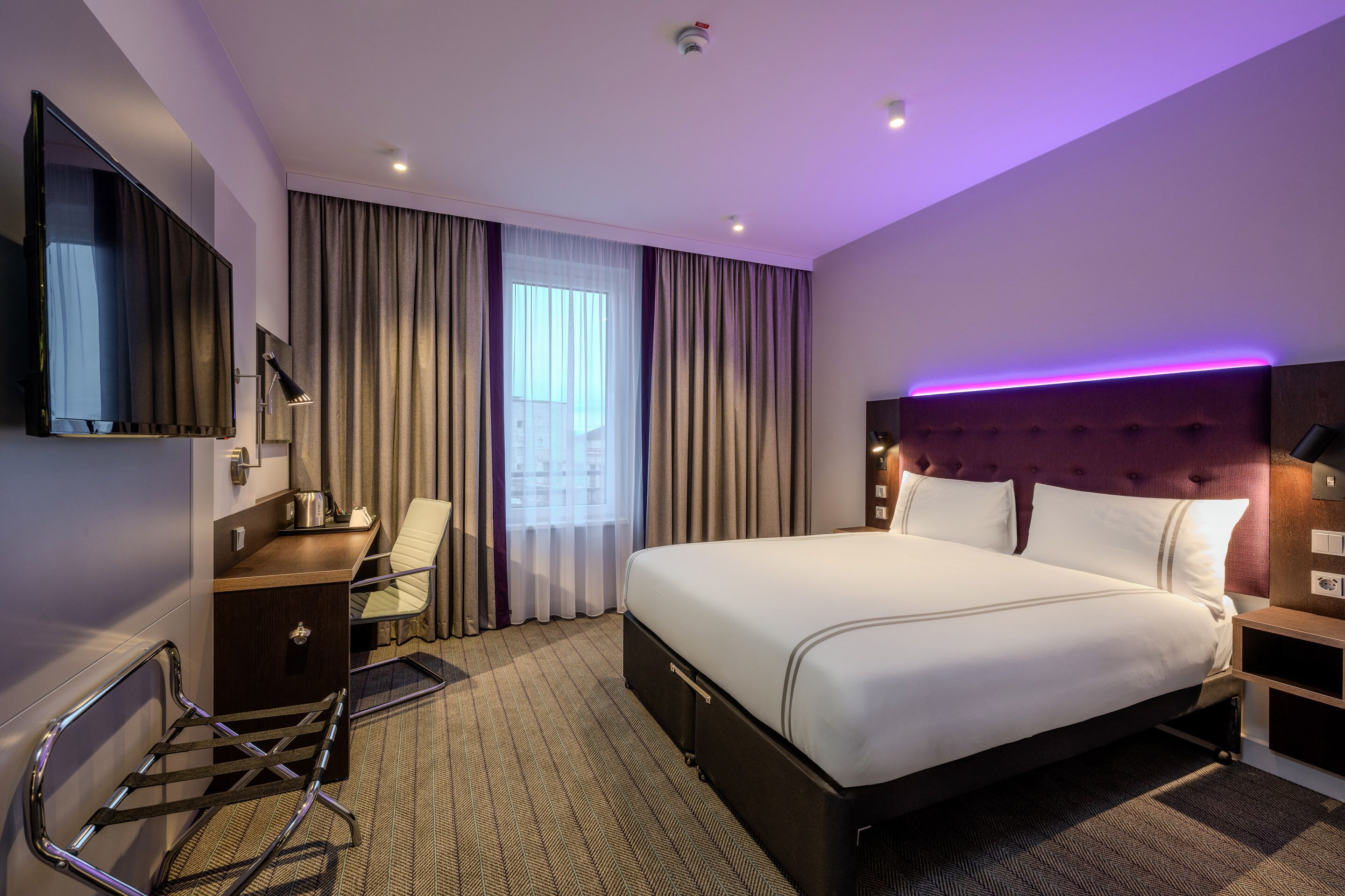A hotel room with a bed, a desk, a chair, a TV, and a window with curtains.