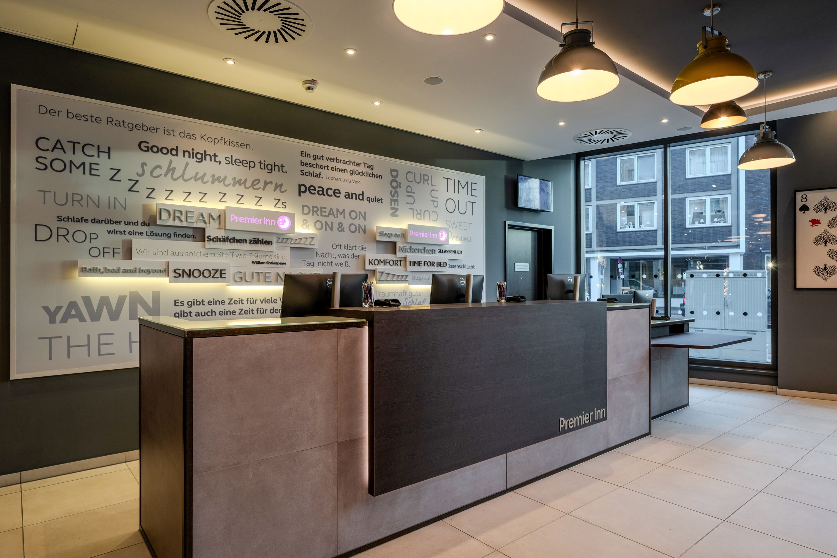 A hotel reception desk with a large wall sign displaying various phrases in German, a modern interior design, and a view of the outside through large windows.