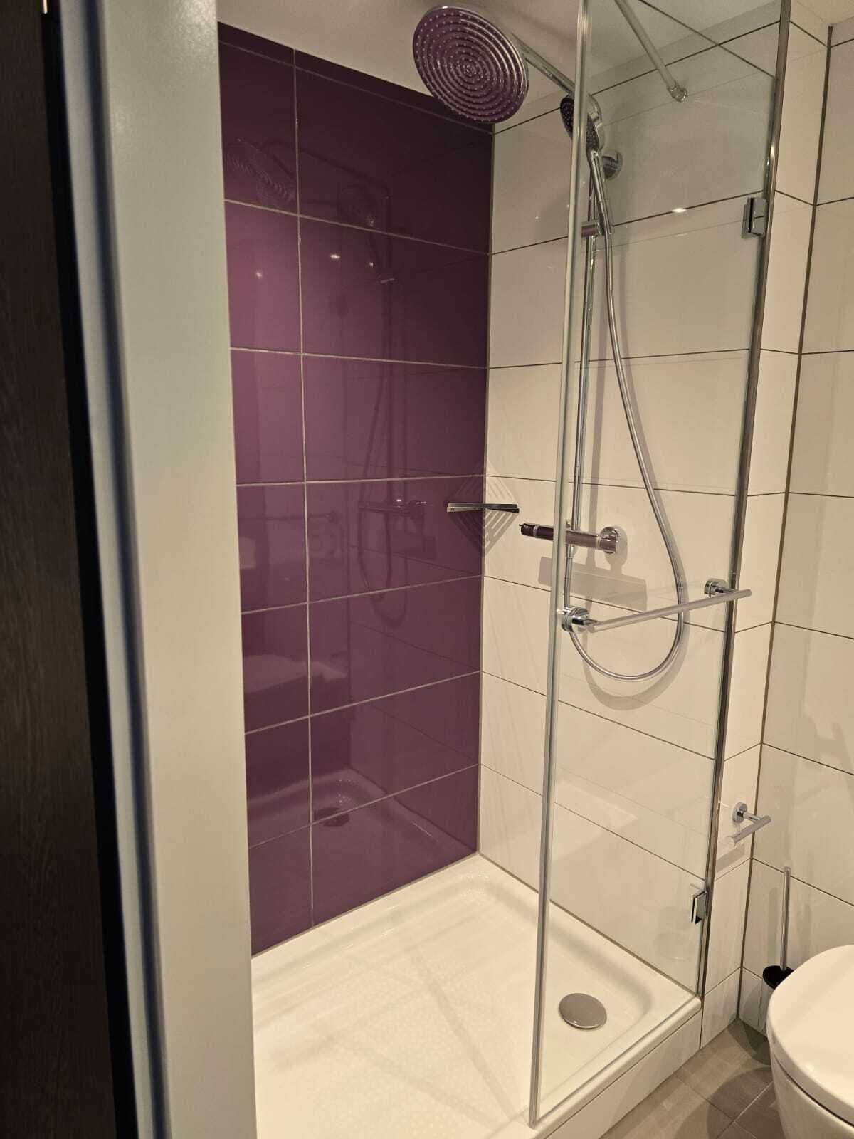 A modern bathroom with a glass shower enclosure, white and purple tiles, and a visible toilet.