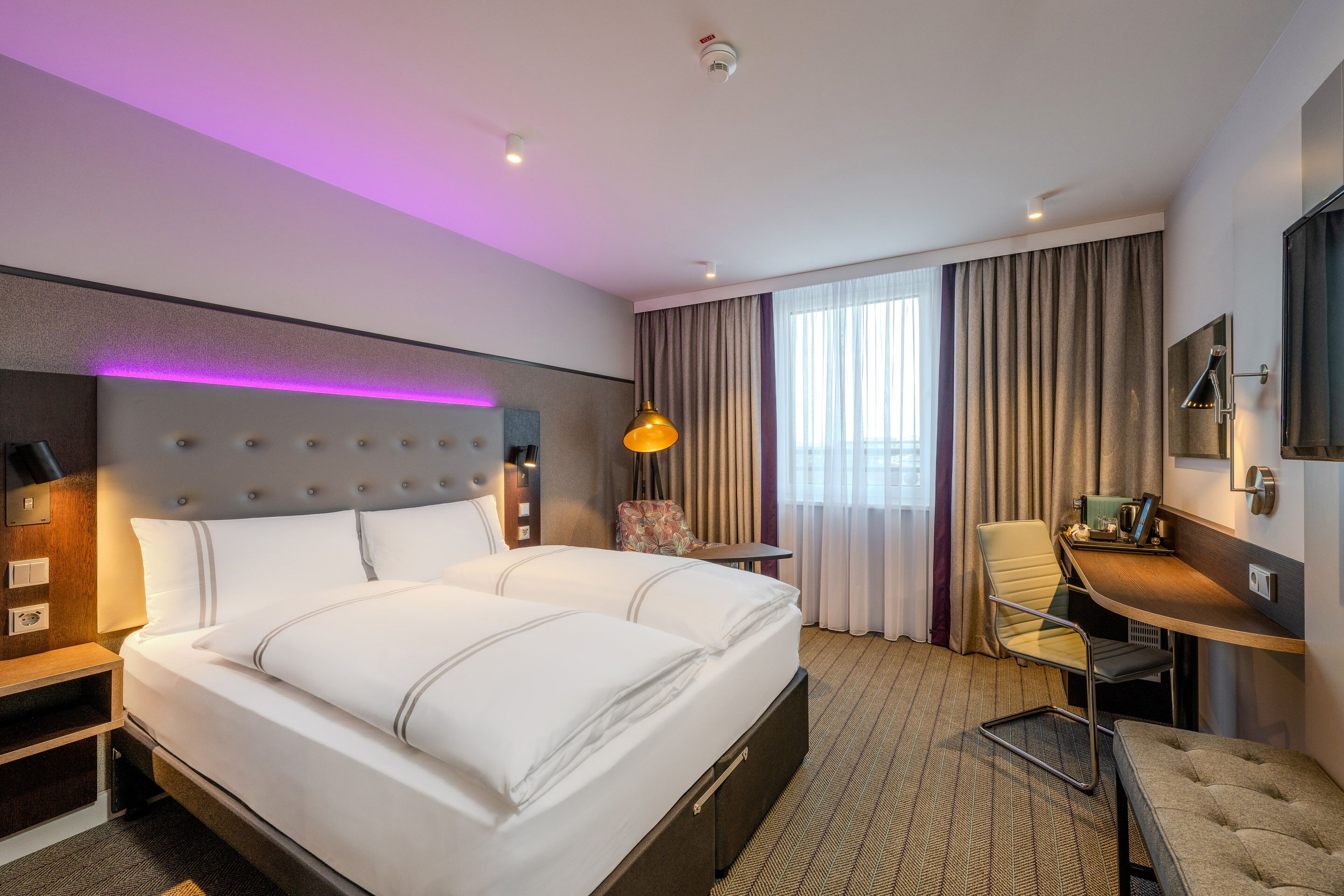 Une chambre d’hôtel avec un grand lit, un bureau, une chaise, une lampe et une fenêtre avec des rideaux.