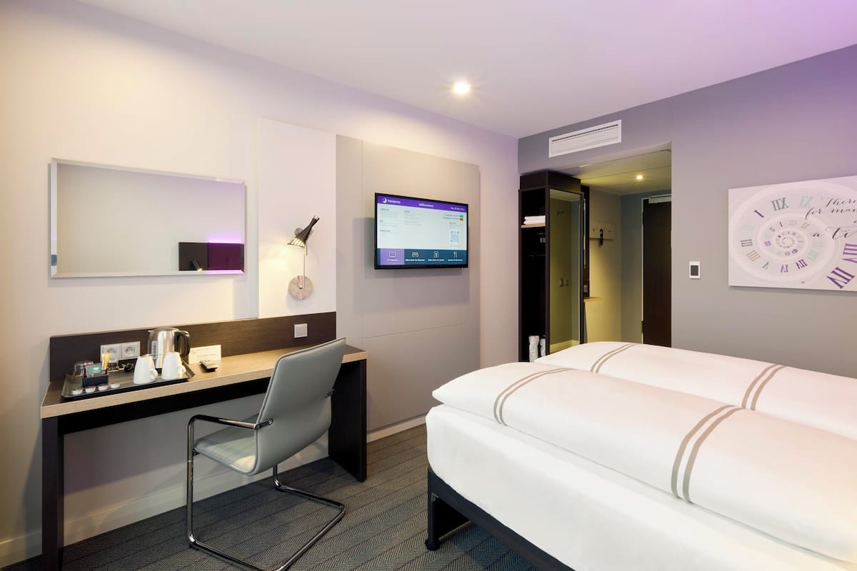 A hotel room with a bed, a desk with a chair, a flat-screen TV, and a wall-mounted clock.