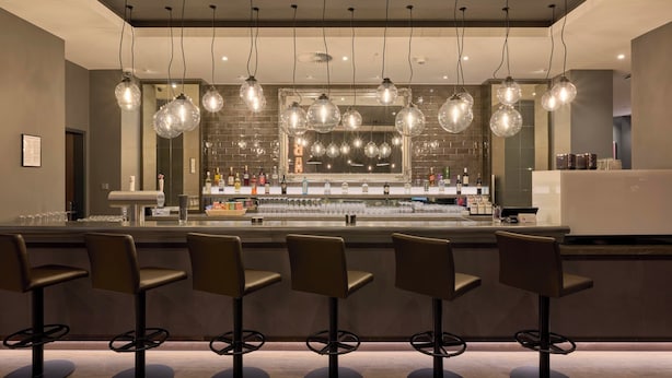 A bar area with multiple stools, hanging light fixtures, and a mirrored wall. - Premier Inn Stuttgart Zuffenhausen (Stuttgart)