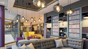 A modern lounge area with a large sectional sofa, round tables, and a bookshelf filled with books. - Premier Inn Stuttgart Zuffenhausen (Stuttgart)