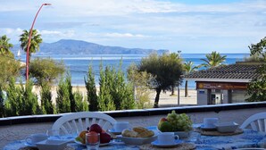 Ett bord dukat med frukostmat, med utsikt över en strand och havet, och i bakgrunden syns en byggnad med skylten "Balesta".