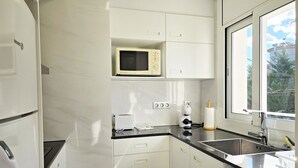 Ett modernt kök med vita skåp, en mikrovågsugn, en brödrost och ett fönster med utsikt över träd och byggnader.