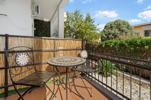 Un balcón con mesa y sillas de metal, una valla de bambú y vistas al entorno, con vegetación y edificios.