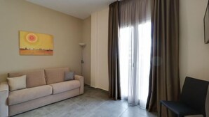 Ein Wohnzimmer mit einem beigefarbenen Sofa, einem schwarzen Sessel, einem Bild an der Wand und einem großen Fenster mit Vorhängen.