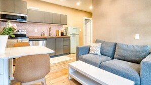 Une cuisine moderne avec des meubles gris, un plan de travail blanc et un four encastré. Le salon est aménagé avec un canapé gris doté d’un coussin blanc et une table basse blanche.