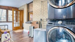 Un espace buanderie comprenant deux machines à laver superposées, un radiateur et une porte en bois donnant sur un espace extérieur.