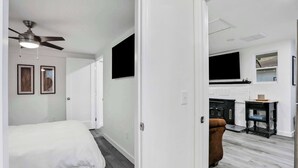 Ein modernes Schlafzimmer mit einem Bett, einem Deckenventilator und einer Tür zu einem Wohnbereich mit Kamin und Fernseher.