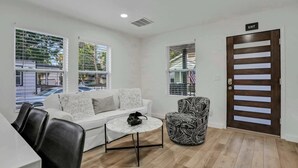 Una sala de estar moderna con un sofá blanco, un sillón estampado, una mesita redonda y una puerta de madera con el letrero 'EXIT'.