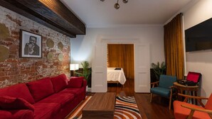 Ein gemütliches Wohnzimmer mit einem roten Sofa, einem Holztisch und einem blauen Sessel. Eine Ziegelwand mit einem gerahmten Bild, ein Bett mit weißer Bettdecke und ein Fernseher an der Wand.