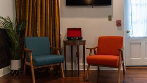 Ein Zimmer mit zwei Stühlen, einem kleinen Tisch mit einer roten Kiste, einer Topfpflanze, einem Fernseher und einem Rauchmelder.