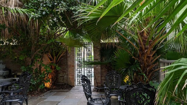Eine Terrasse im Freien mit Steinböden, umgeben von üppiger Bepflanzung und verzierten Metallstühlen.