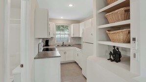 A modern kitchen with white cabinetry, a stainless steel countertop, and a built-in oven. There are wicker baskets on a shelf and a decorative black sculpture on the floor.