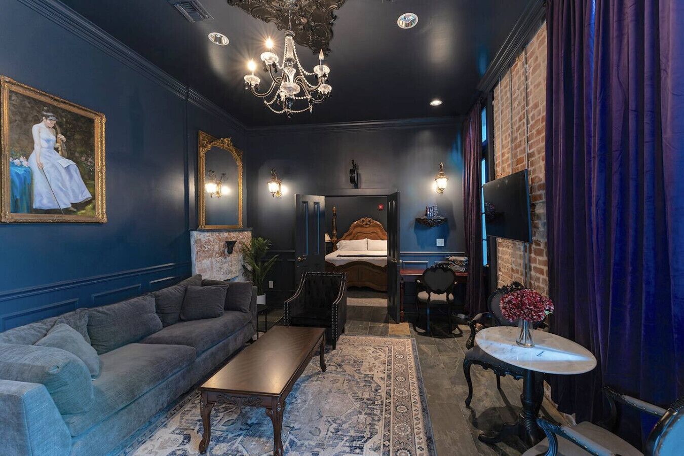 A room with a dark blue wall, a large sofa, a small table, a chandelier, and a bed with a headboard.