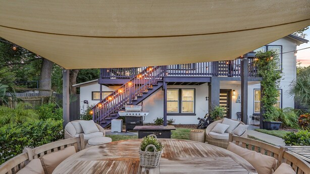 Een overdekt terras met een ronde houten tafel en zachte zitplaatsen, een barbecue en een trap naar een balkon op de tweede verdieping.