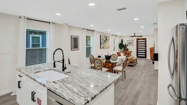Una cocina moderna con encimera de mármol, armarios blancos y un lavamanos integrado. Hay una zona de comedor con una mesa y sillas de vidrio, y un ventilador de techo.