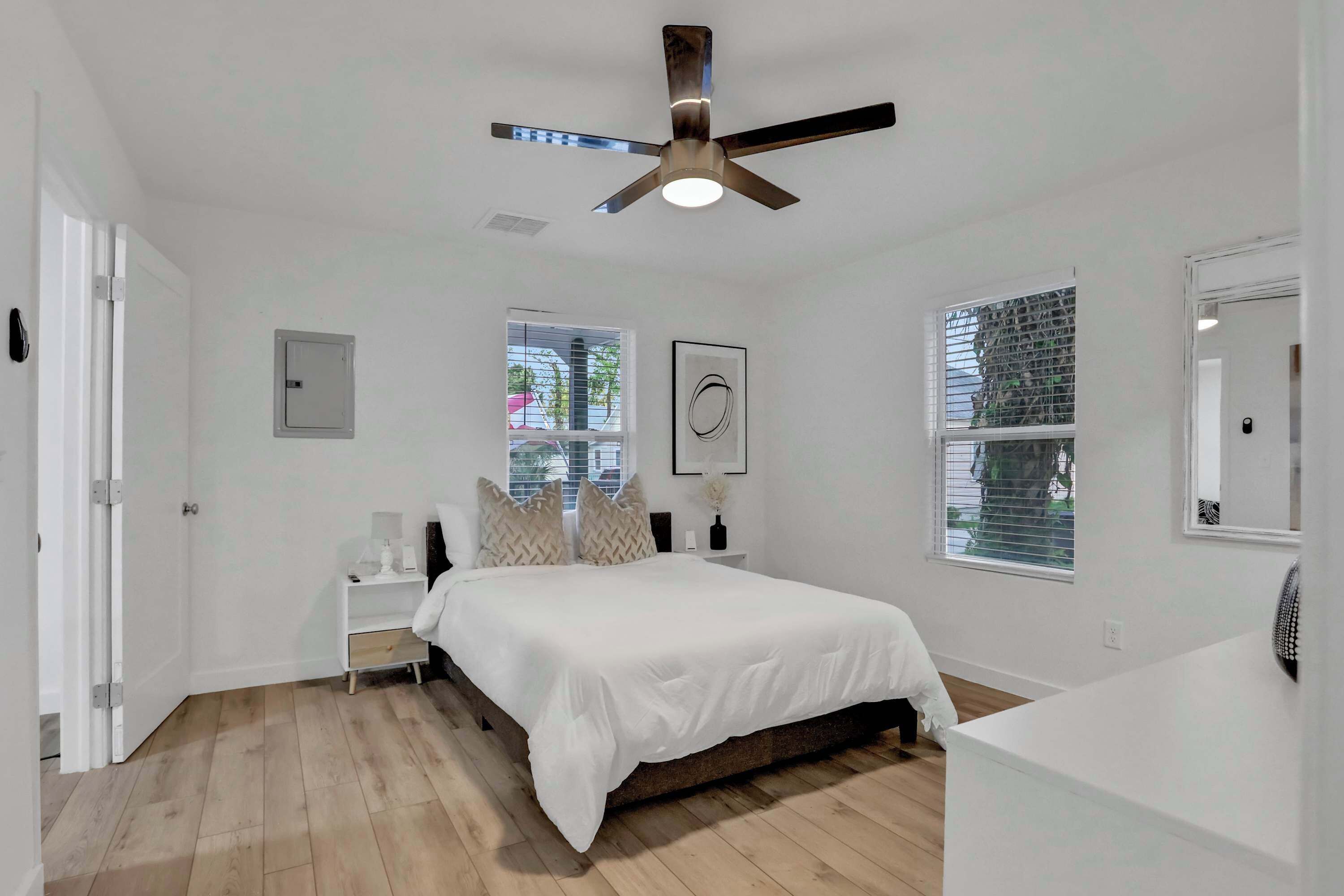 Un dormitorio con cama, mesitas de noche, ventilador de techo y dos ventanas.