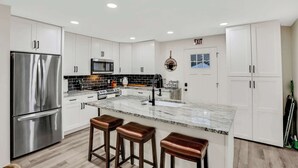 Una cocina moderna con armarios blancos, electrodomésticos de acero inoxidable, una isla de mármol con taburetes y vistas al exterior.