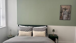 A bedroom with a bed, two bedside tables, a nightstand, a window, and a wall-mounted artwork. - Stunning 3 Bed Apartment By Urban Roost (Worthing)