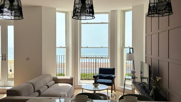 A modern living room with a sofa, dining table, and chairs. Large windows offer a view of the beach. - Stunning 3 Bed Apartment By Urban Roost (Worthing)
