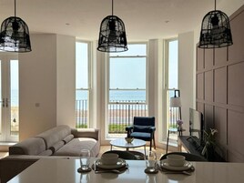 Una sala de estar moderna con sofá, mesa de comedor y sillas. Amplias ventanas ofrecen vistas a la playa.