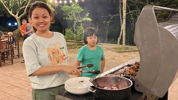 Un barbecue avec des brochettes, une casserole et des légumes coupés en tranches, le tout posé sur une table.
