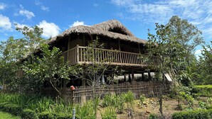 A traditional thatched-roof house with a wooden balcony, surrounded by lush greenery and a clear blue sky.