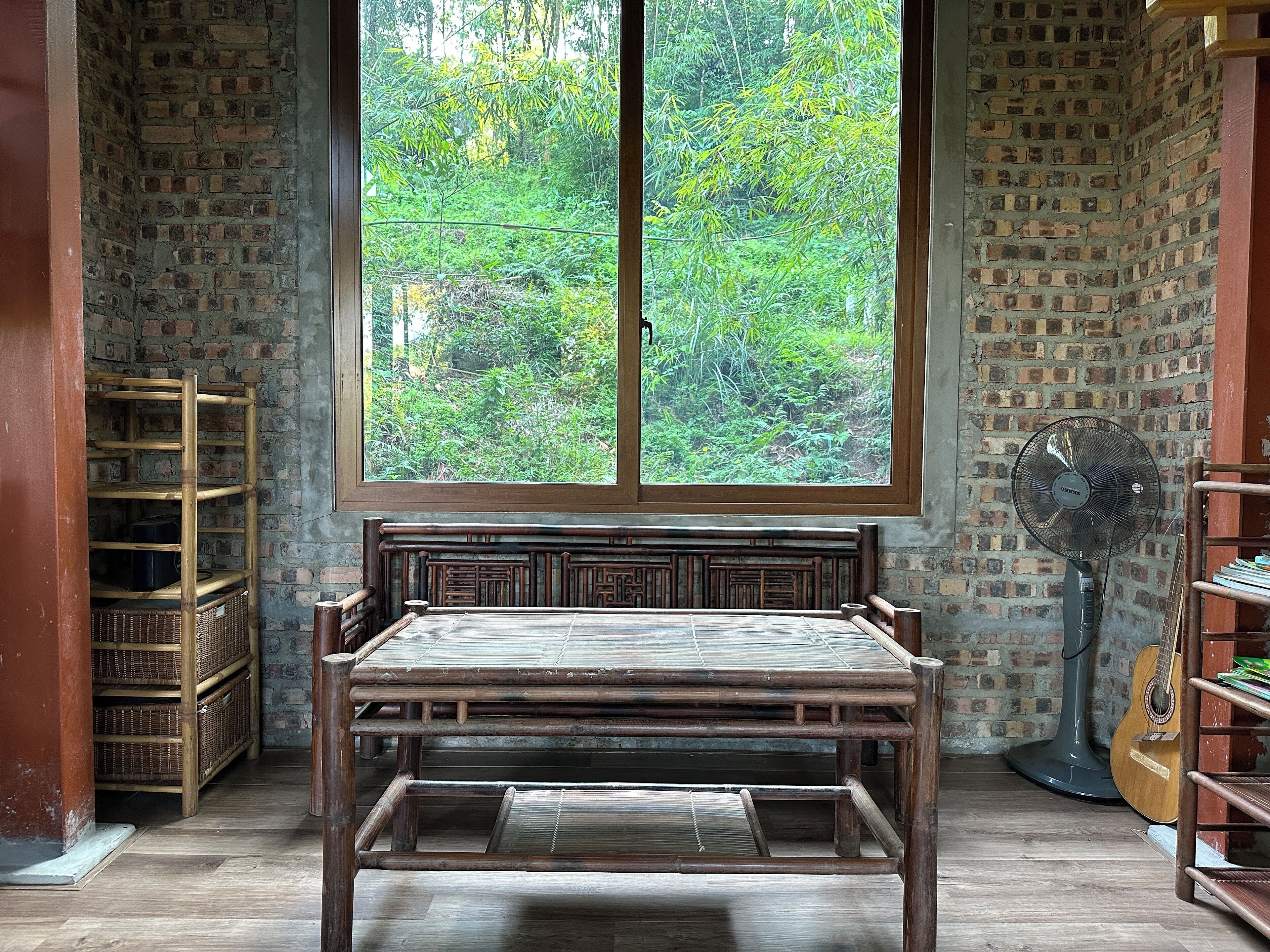 Una habitación con una cama de madera, un ventanal con vistas a zonas verdes, un ventilador y estantes con objetos.