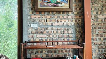 A room with a brick wall, a wooden bench with a red cushion, a framed painting of flowers, and a window with a view of greenery.