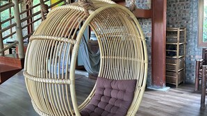 A wicker hanging chair with a cushioned seat, suspended by ropes, in a room with wooden flooring and brick walls.